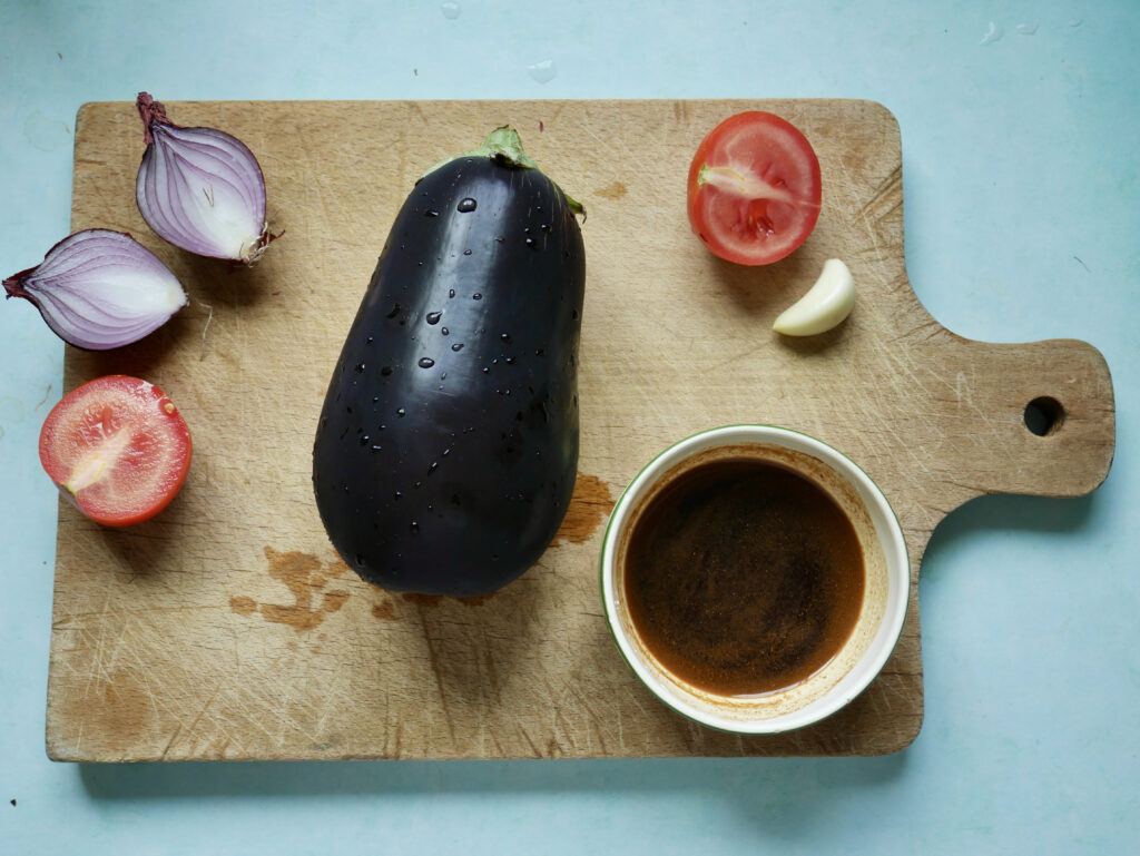 Aubergine, Tomate, Knoblauchzehe, Schälchen mit Soße und Zwiebel auf einem Holz-Schneidebrett