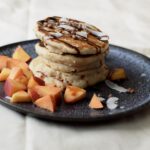 Pancake Stack garniert mit Kokoschips, Dattelsirup und Nektarinenstücken auf einem dunklen Keramikteller